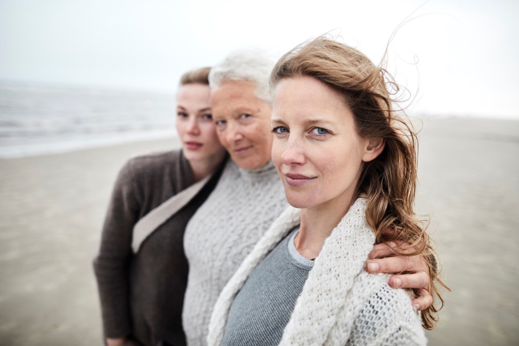 Three women standing astride each other on a beach with arms over each other's shoulders. The woman on the left is younger, in a green shirt with a bag strap over her shoulder. The woman in the middle is older with white hair and a gray sweater. The women on the right in middle-aged with longer, light brown hair. She is wearing a white cardigan with a darker grey crewneck underneath.